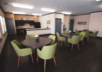 Community room with 3 brown tables each surrounded by green chairs and a kitchen prep area